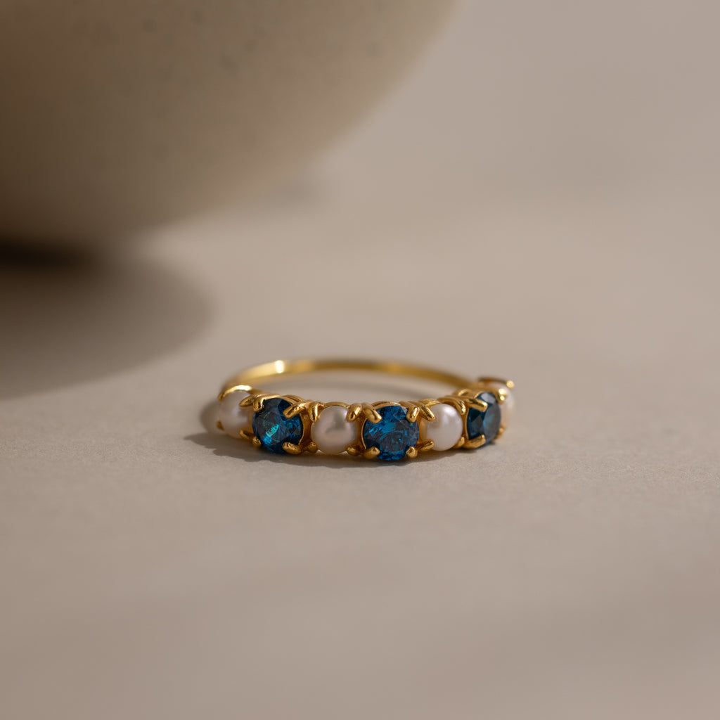 The Blue Zircon & Pearl Lace Ring features alternating blue zircons and white pearls on a gold band, elegantly displayed—ideal as a unique birthstone or pearl ring.