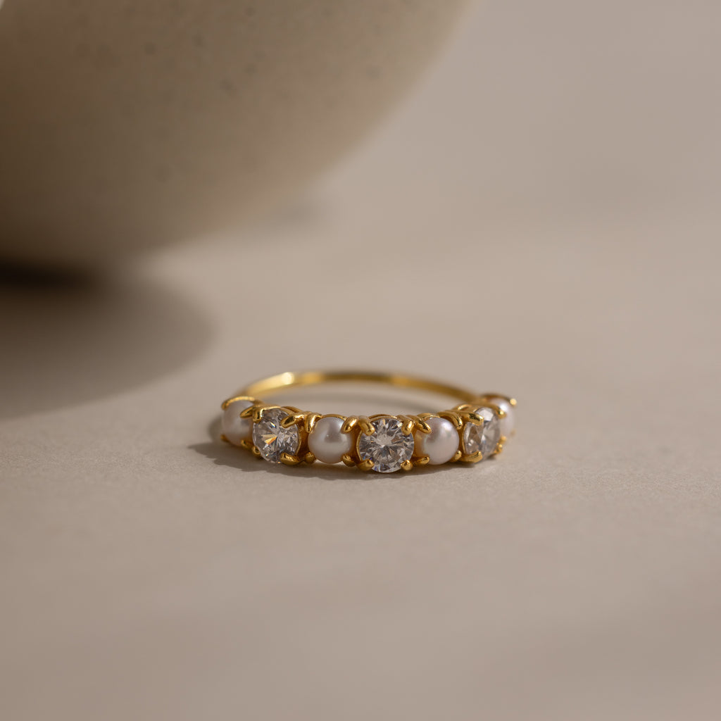 Diamond & Pearl Lace Ring featuring a sleek gold band with white pearls and round clear gemstones, elegantly displayed on a neutral beige surface.