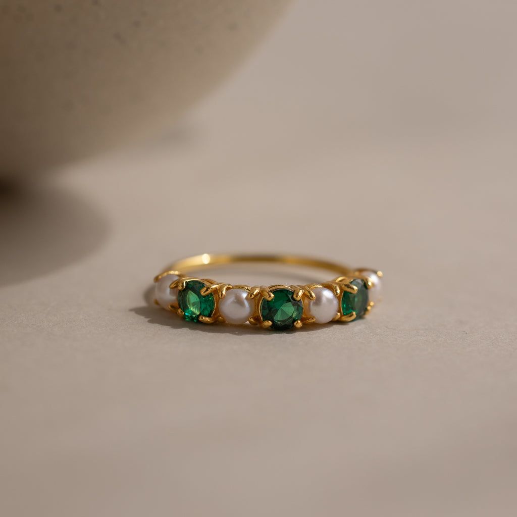 The Emerald & Pearl Lace Ring, a gold band adorned with alternating emeralds and pearls, rests on a light, neutral surface.