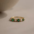 The Emerald & Pearl Lace Ring, a gold band adorned with alternating emeralds and pearls, rests on a light, neutral surface.