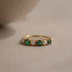 The Emerald & Pearl Lace Ring, a gold band adorned with alternating emeralds and pearls, rests on a light, neutral surface.