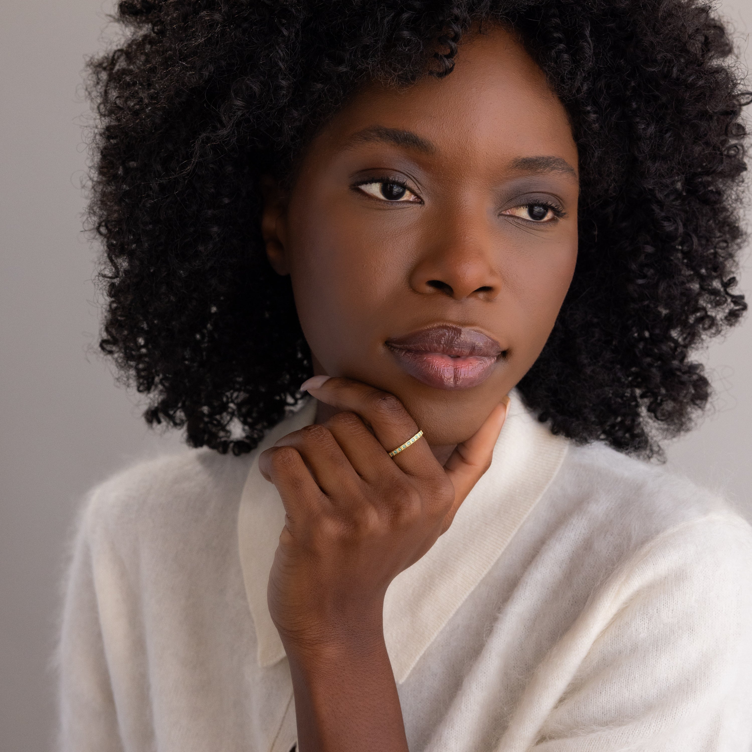 A woman with curly hair in a white sweater rests her chin on her hand, thoughtfully admiring the Art Deco Aquamarine Eternity Ring on her finger.