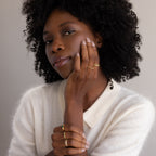 A woman with curly hair in a white sweater poses with her hands on her face, elegantly showcasing gold rings, including the Art Deco Emerald Eternity Ring and a dazzling birthstone ring.