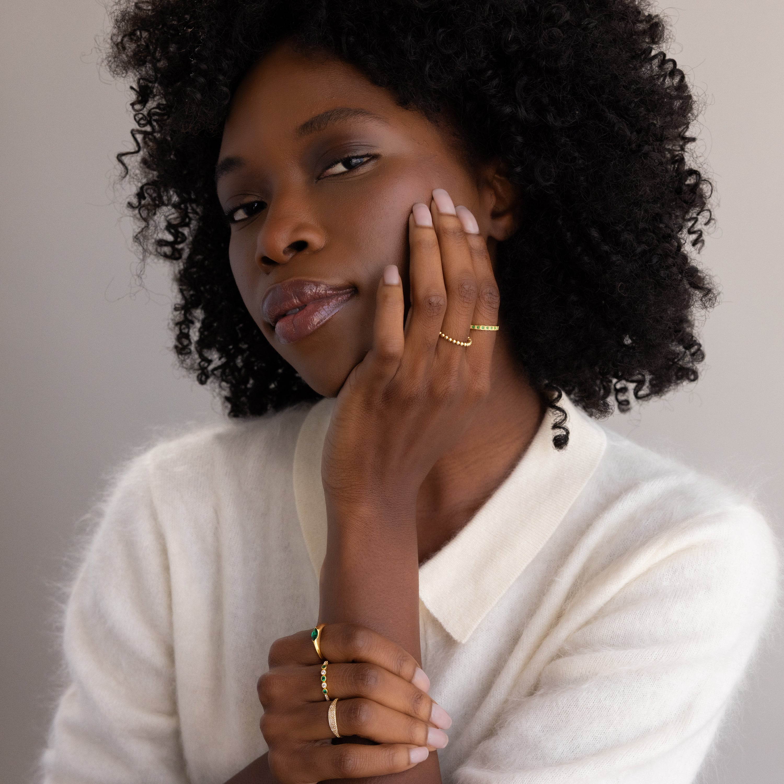 A woman with curly hair in a white sweater poses with her hands on her face, elegantly showcasing gold rings, including the Art Deco Emerald Eternity Ring and a dazzling birthstone ring.