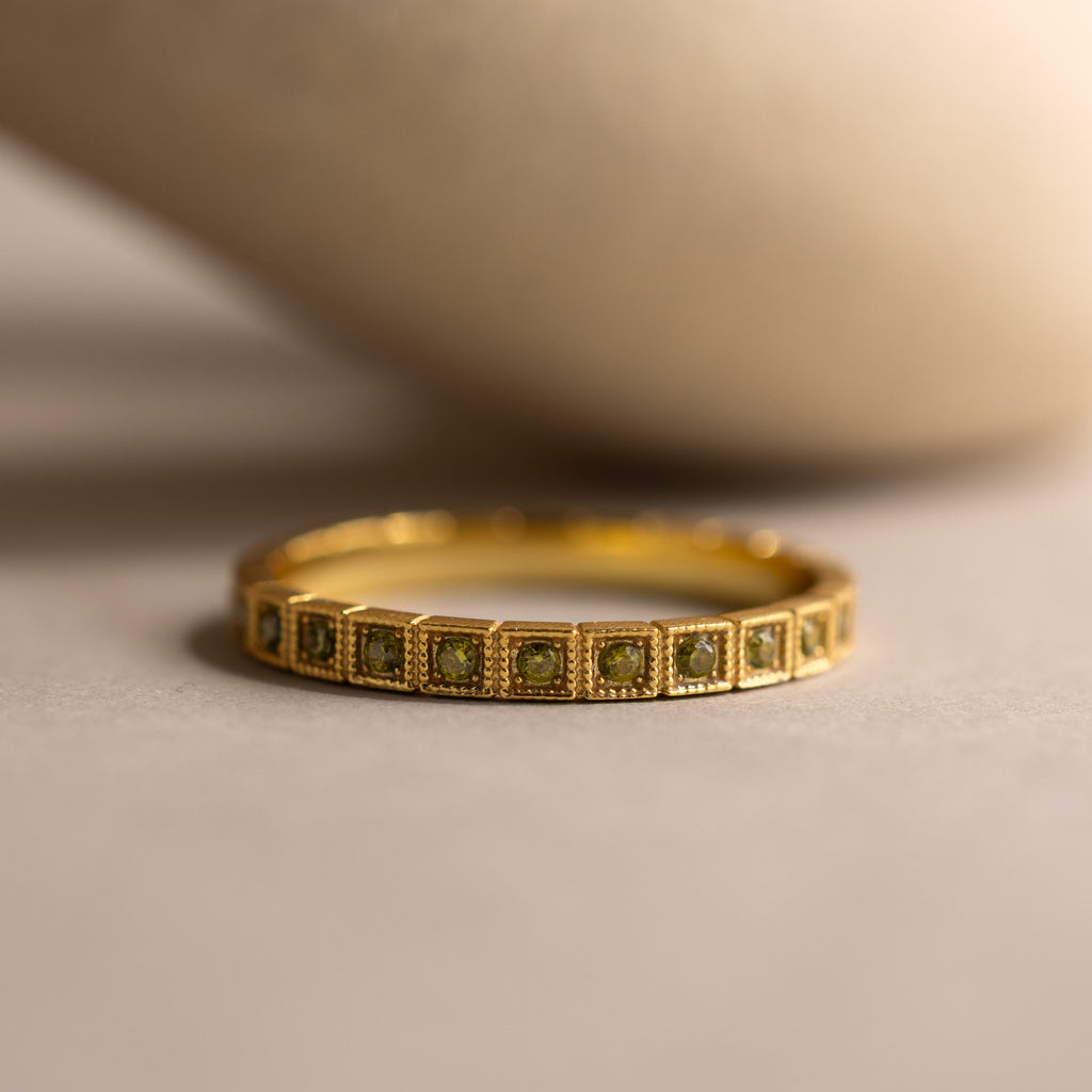 The Art Deco Peridot Eternity Ring features square green peridot gemstones set in a row on a neutral surface—a striking vintage-inspired jewelry piece.
