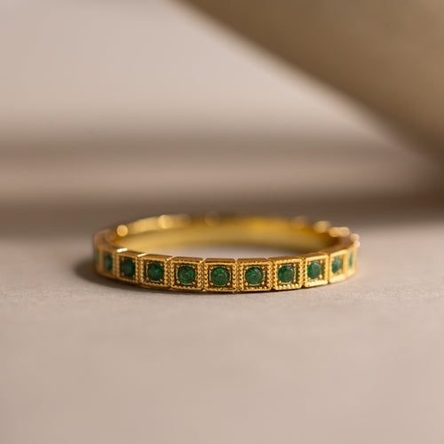 The Art Deco Emerald Eternity Ring showcases a row of small square emeralds in a vintage bezel setting, displayed on a neutral surface.