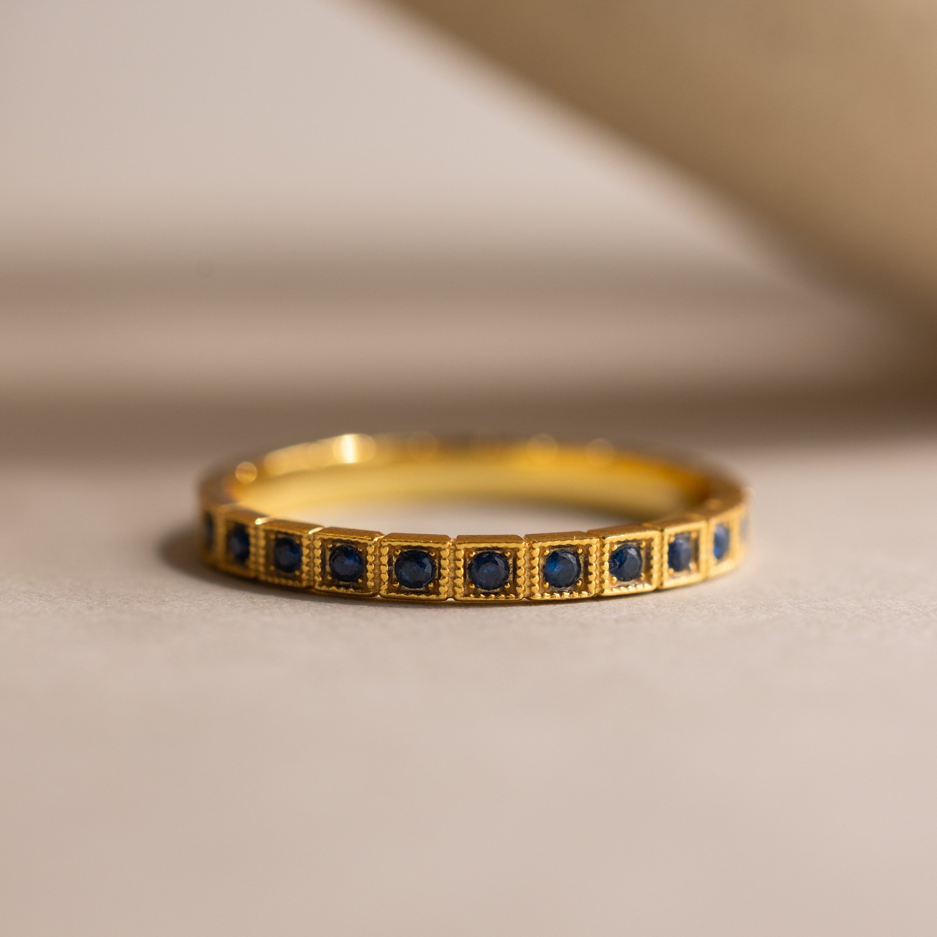 The Art Deco Sapphire Eternity Ring features small blue sapphires set in a row, showcased on a neutral, softly lit surface.