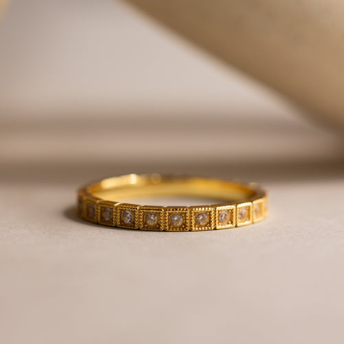 The Art Deco Diamond Eternity Ring features a gold band with small square-shaped diamond settings, showcased on a neutral surface.