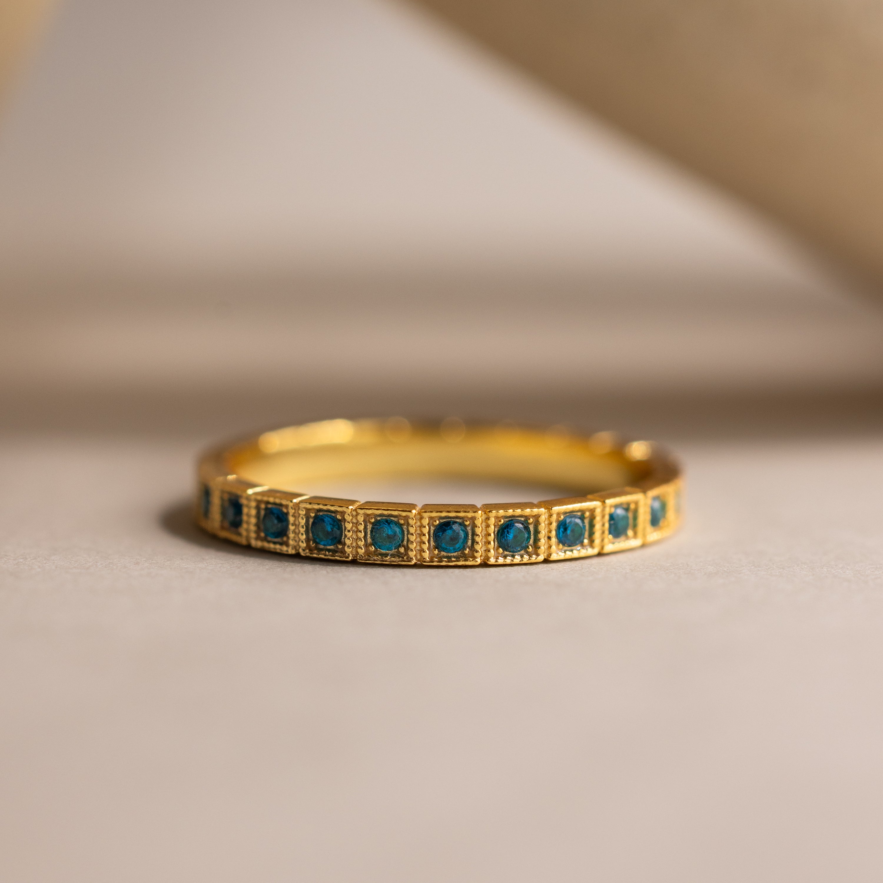 The Art Deco Blue Zircon Eternity Ring features vintage-inspired gold design with small square blue zircons set around the band, displayed on a neutral surface.