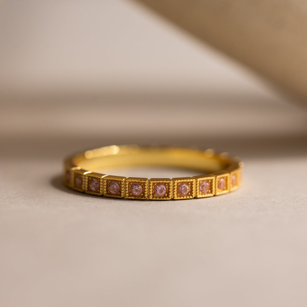 The Art Deco Pink Tourmaline Eternity Ring features square pink tourmalines in a row, set in gold with vintage bezel-milgrain details—a timeless and elegant birthstone ring displayed on a neutral background.
