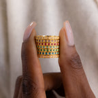 A hand holds the Art Deco Emerald Eternity Ring, a wide gold band set with red, green, and white stones in a grid pattern.