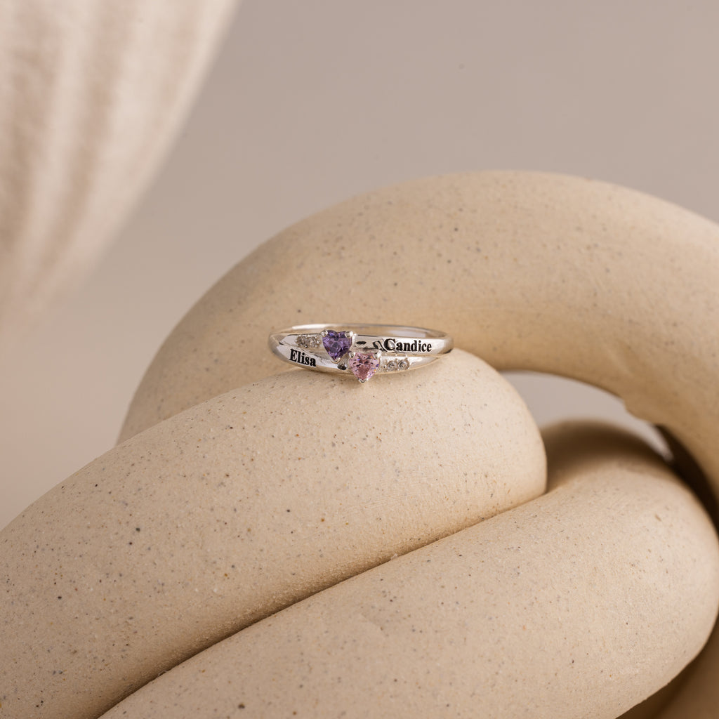 The Toi et Moi Interlocking Name Ring, featuring two heart-shaped birthstones and engraved names, rests on a beige, twisted sculpture.