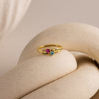 Toi et Moi Interlocking Name Ring in 18K Gold features two heart-shaped gemstones and engraved names, showcased on a textured, cream-colored surface.