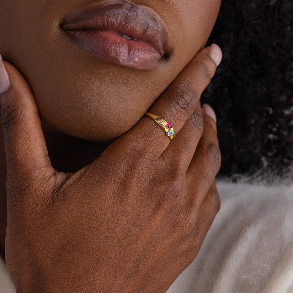 A hand with a Toi et Moi Interlocking Name Ring in 18K Gold, personalized with birthstones, touches a person’s chin.