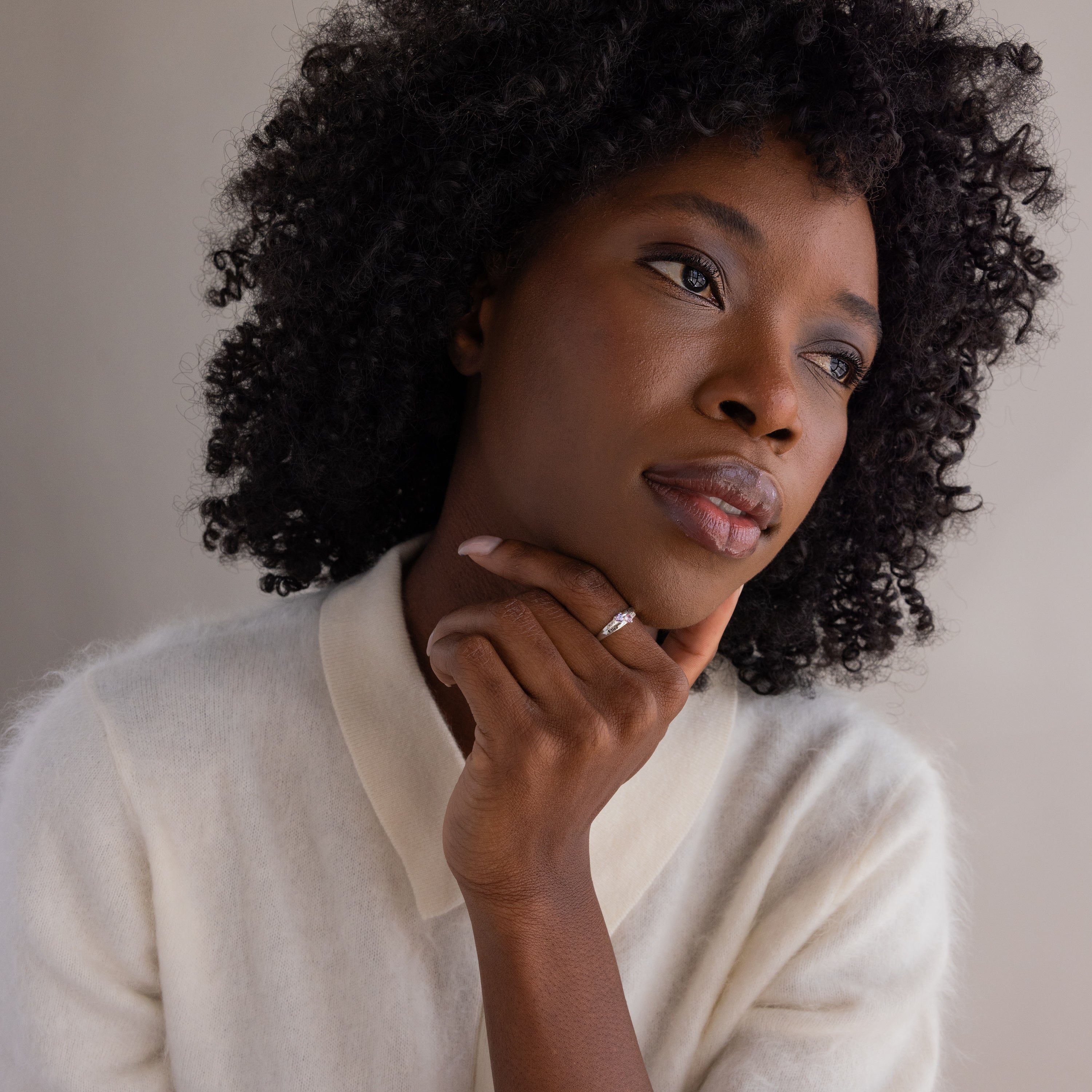 A woman with curly hair in a white sweater looks thoughtful, resting her chin on her hand and wearing the Toi et Moi Interlocking Name Ring in Sterling Silver.