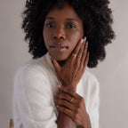 A woman with curly hair in a white sweater touches her face, displaying the Toi et Moi Interlocking Name Ring in 18K Gold as she gazes at the camera.