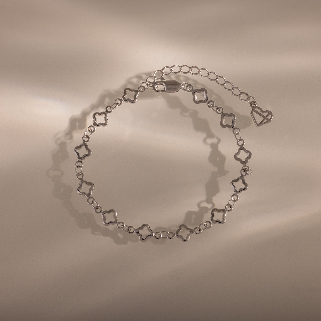 The Clover Station Bracelet in Silver features delicate clover-shaped links and a heart charm, elegantly displayed on a beige surface.