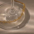 The Clover Station Bracelet in 18K Gold, featuring delicate floral links, is draped around the base of a wine glass on a light surface.