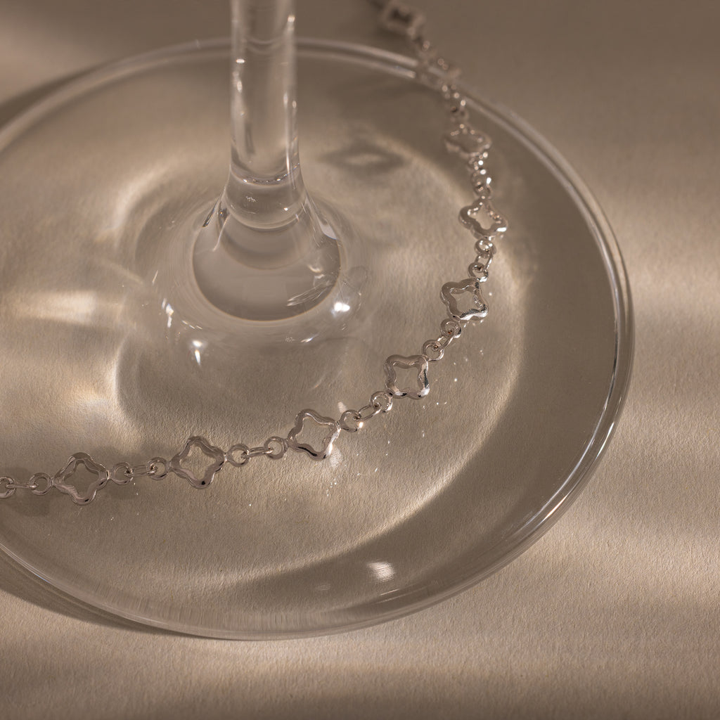 The Clover Station Bracelet in Silver, with its star-shaped links, is elegantly draped around the base of a glass on a softly lit beige surface.