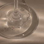 The Clover Station Bracelet in Silver, with its star-shaped links, is elegantly draped around the base of a glass on a softly lit beige surface.