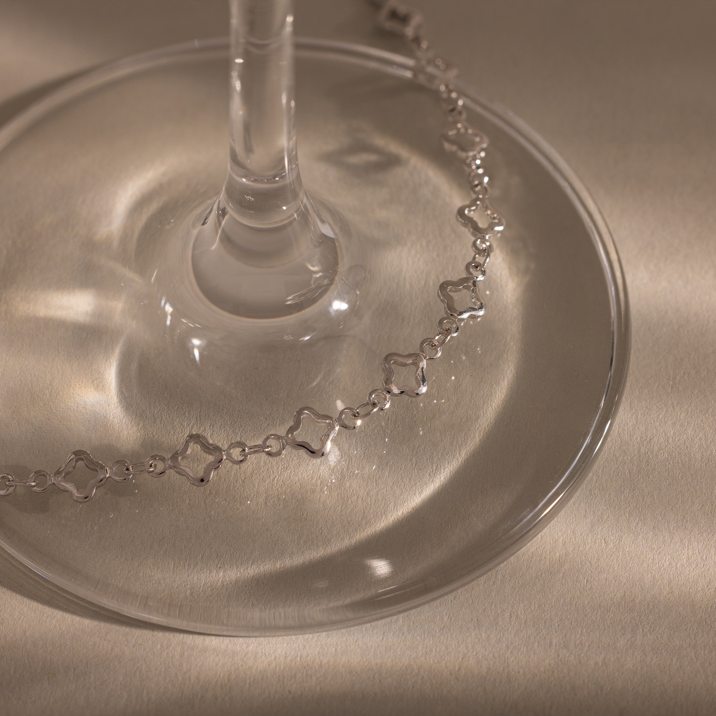 The Clover Station Bracelet in Silver, with its star-shaped links, is elegantly draped around the base of a glass on a softly lit beige surface.