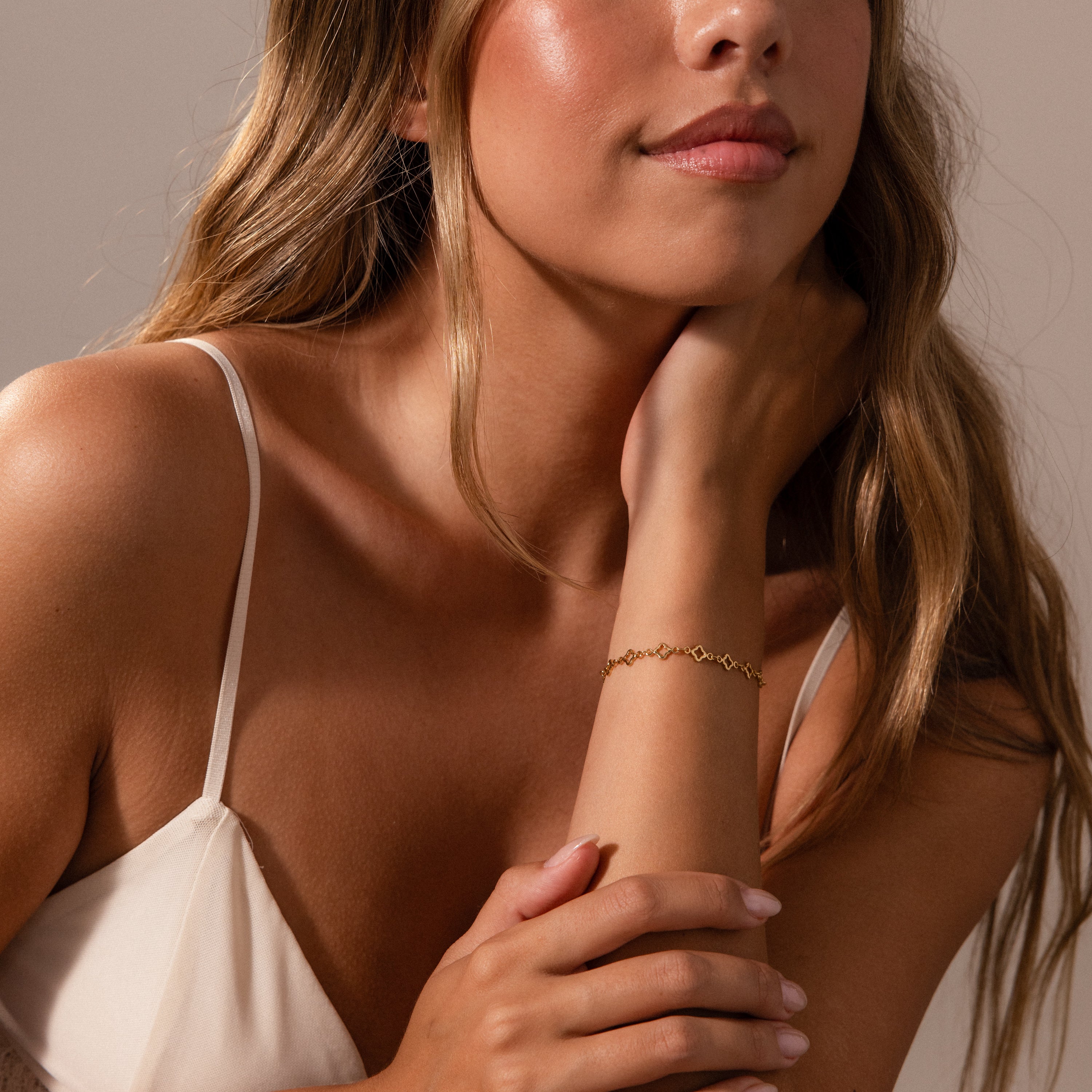 Woman with long wavy hair wearing a white top and the Clover Station Bracelet in 18K Gold, resting her hand on her neck.