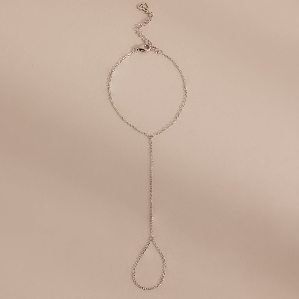 Minimalist silver hand chain bracelet with delicate links and a teardrop-shaped loop, laid flat against a beige background.