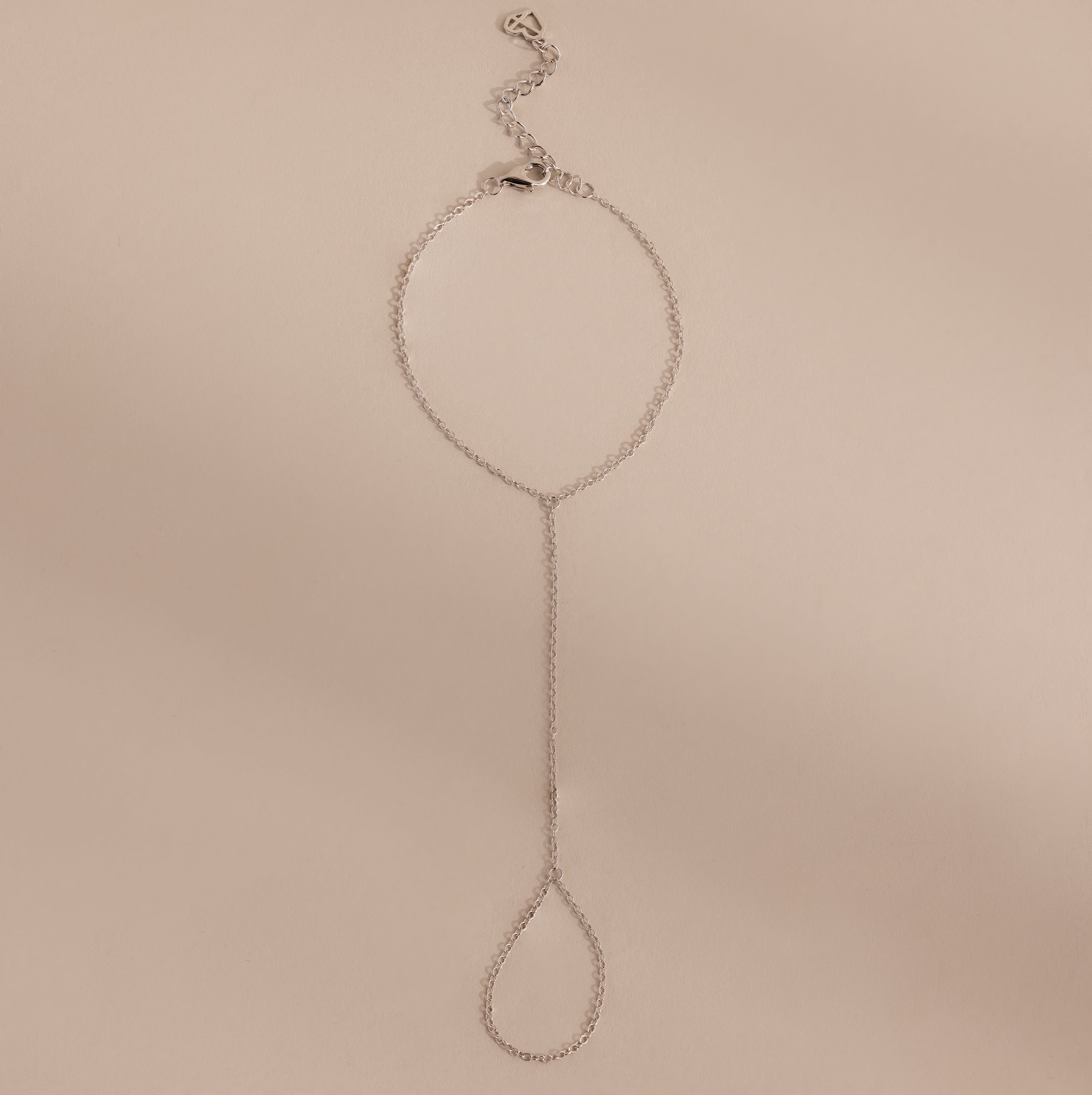 Minimalist silver hand chain bracelet with delicate links and a teardrop-shaped loop, laid flat against a beige background.