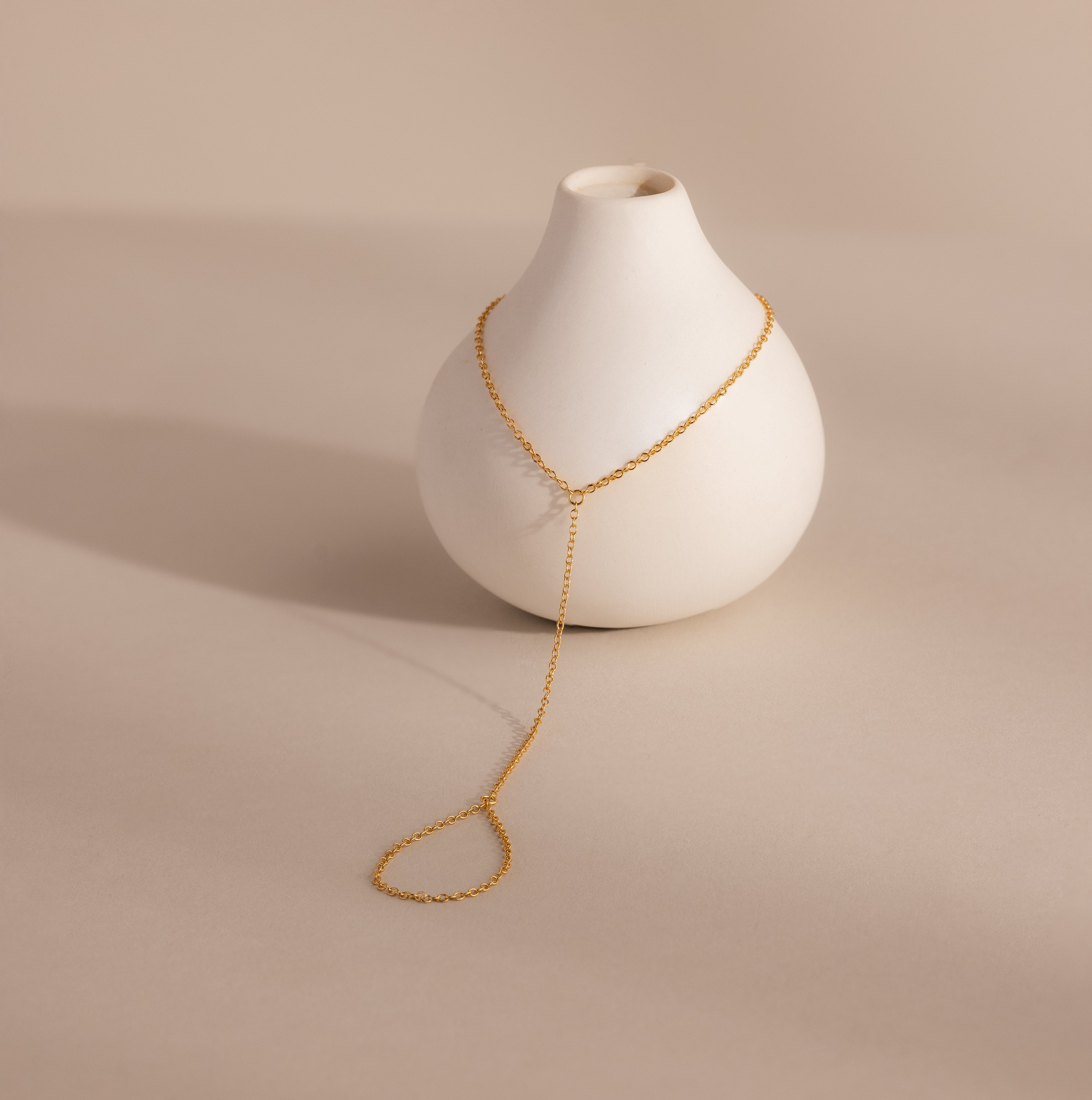 Delicate gold hand chain bracelet elegantly draped over a small matte white ceramic vase, casting a soft shadow.