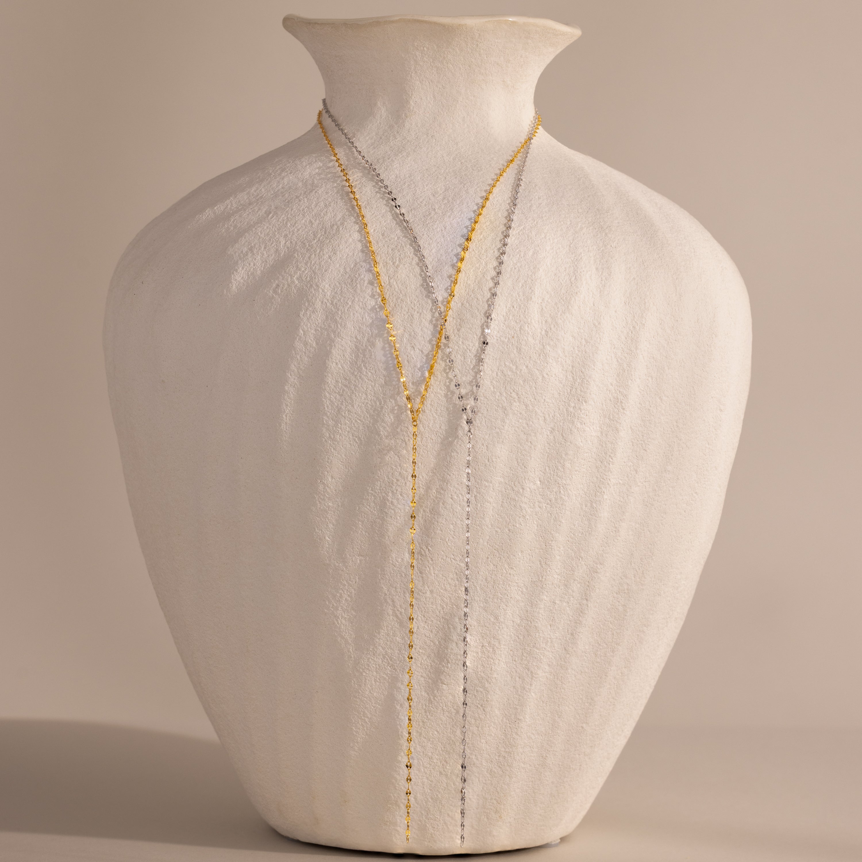 A white textured vase displays two delicate necklaces, including the Mirror Chain Lariat Necklace with a reflective finish, elegantly draped over the front.