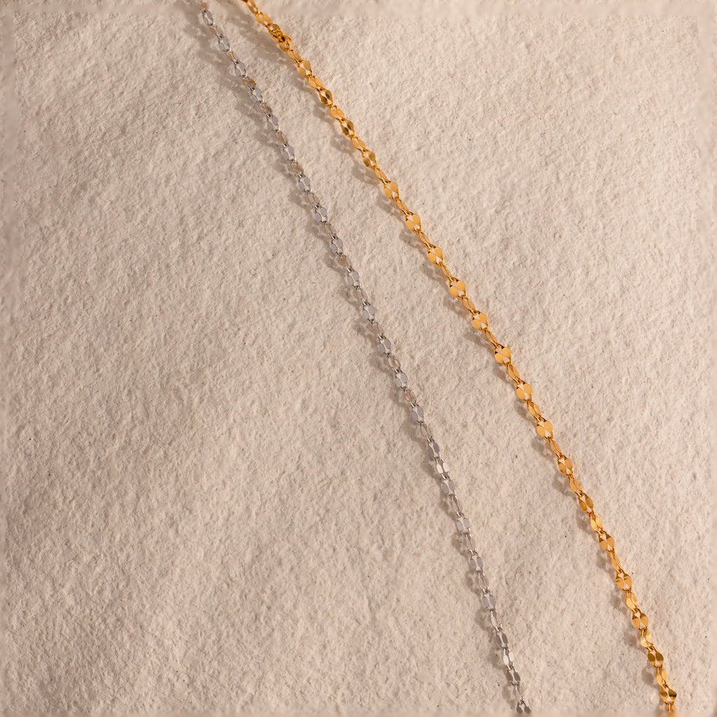 An 18K gold Mirror Chain Lariat Necklace is laid out diagonally on a textured beige surface, highlighting its sleek, reflective finish.