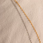 An 18K gold Mirror Chain Lariat Necklace is laid out diagonally on a textured beige surface, highlighting its sleek, reflective finish.