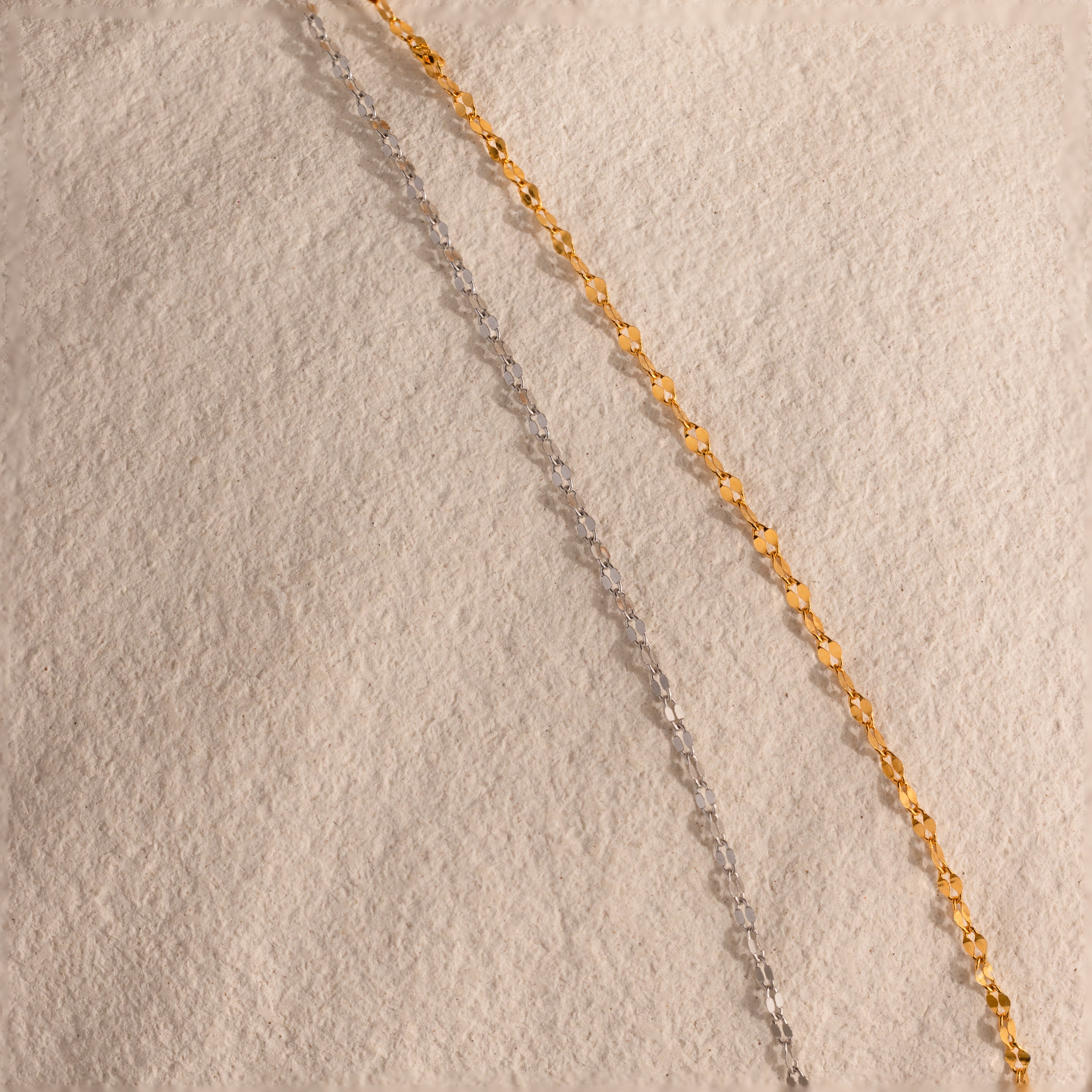 An 18K gold Mirror Chain Lariat Necklace is laid out diagonally on a textured beige surface, highlighting its sleek, reflective finish.
