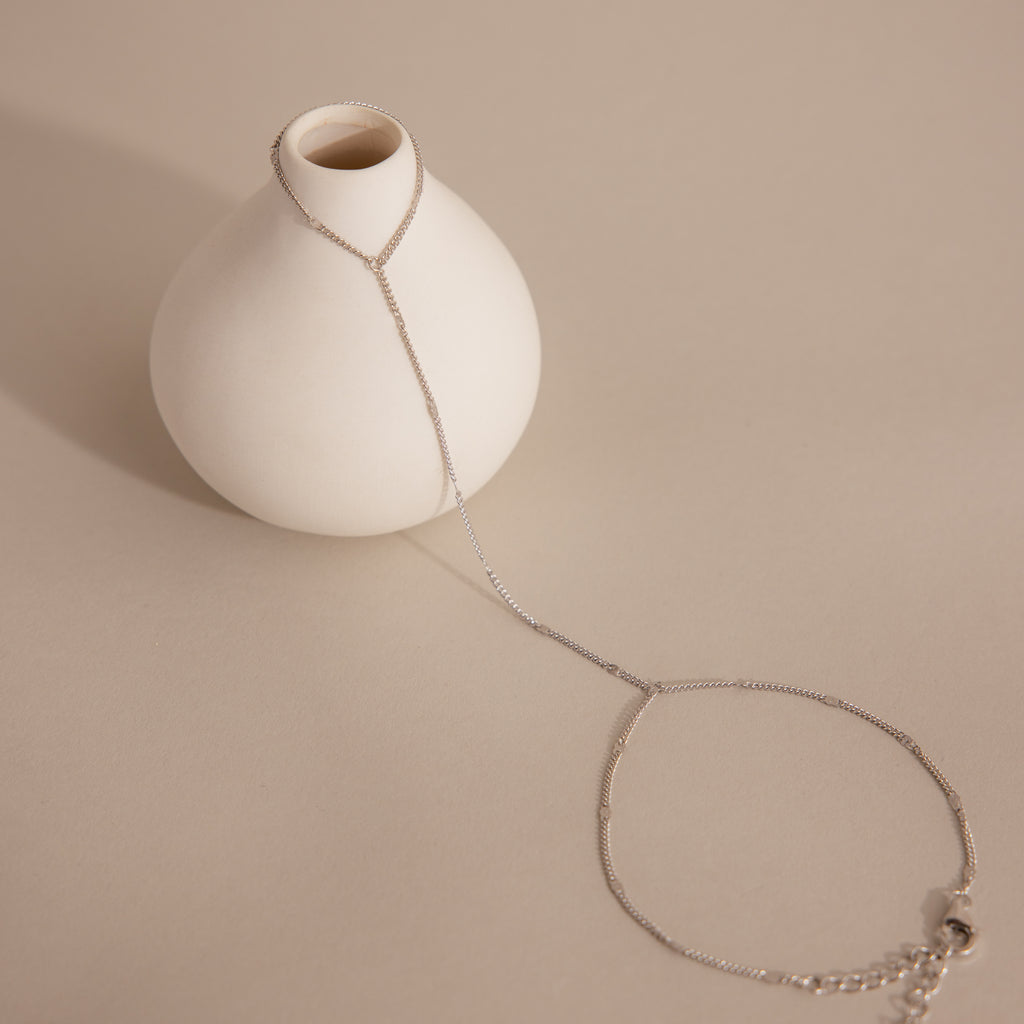 Sterling silver Curb Hand Chain Bracelet with the ring chain resting against a white ceramic prop, while the bracelet is laid on a neutral-tone surface.