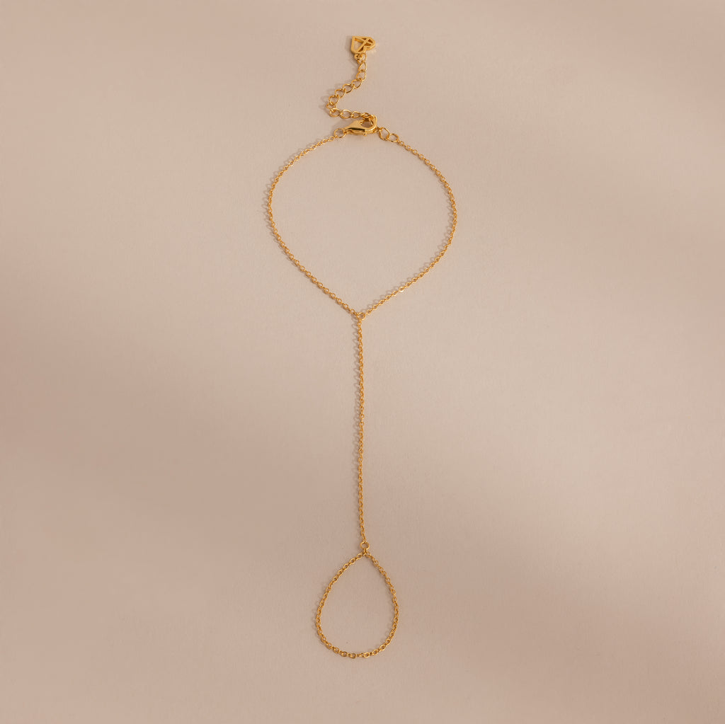 Minimalist gold hand chain bracelet with delicate links and a teardrop-shaped loop, laid flat against a beige background.