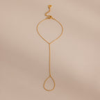 Minimalist gold hand chain bracelet with delicate links and a teardrop-shaped loop, laid flat against a beige background.