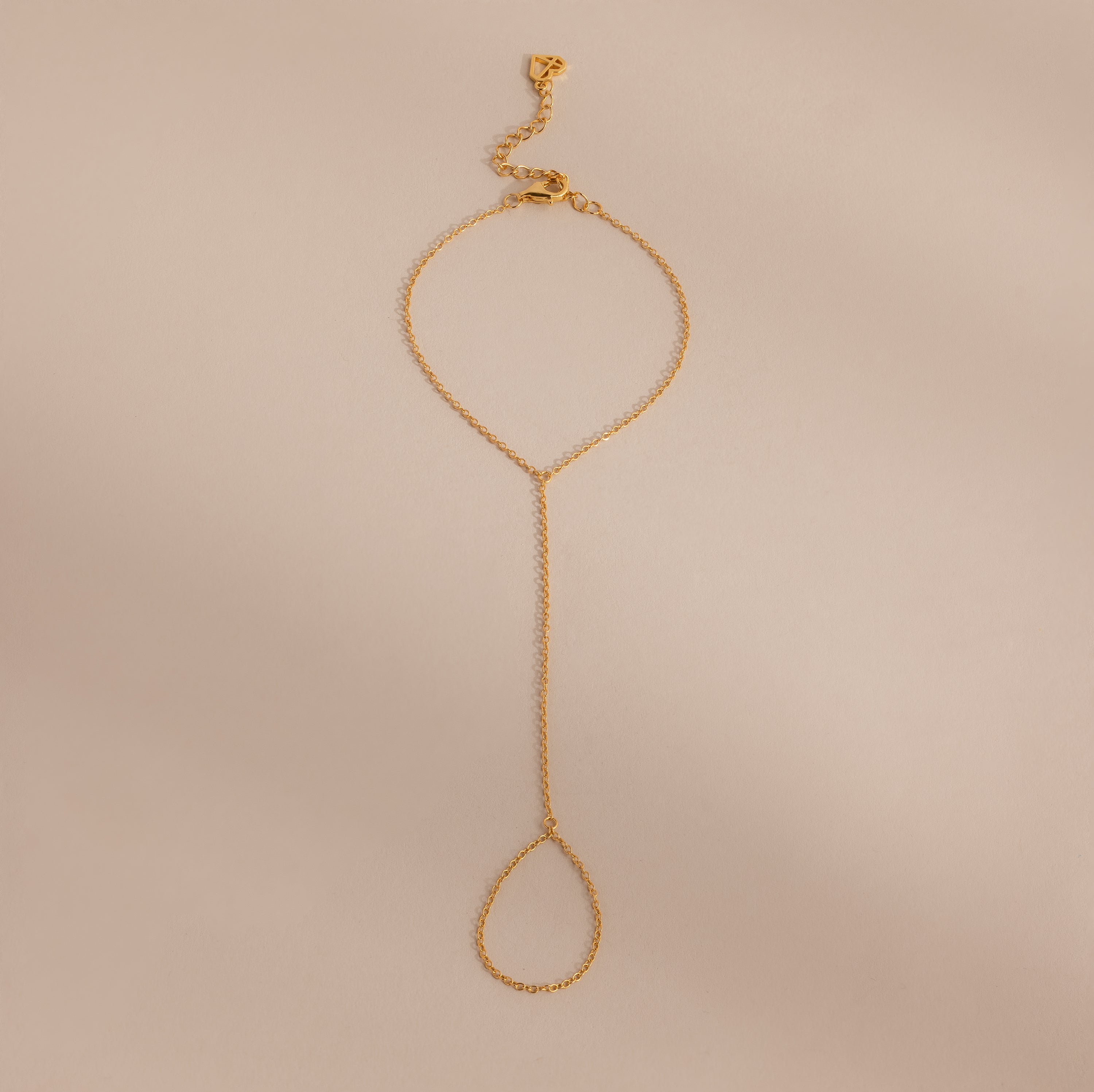 Minimalist gold hand chain bracelet with delicate links and a teardrop-shaped loop, laid flat against a beige background.