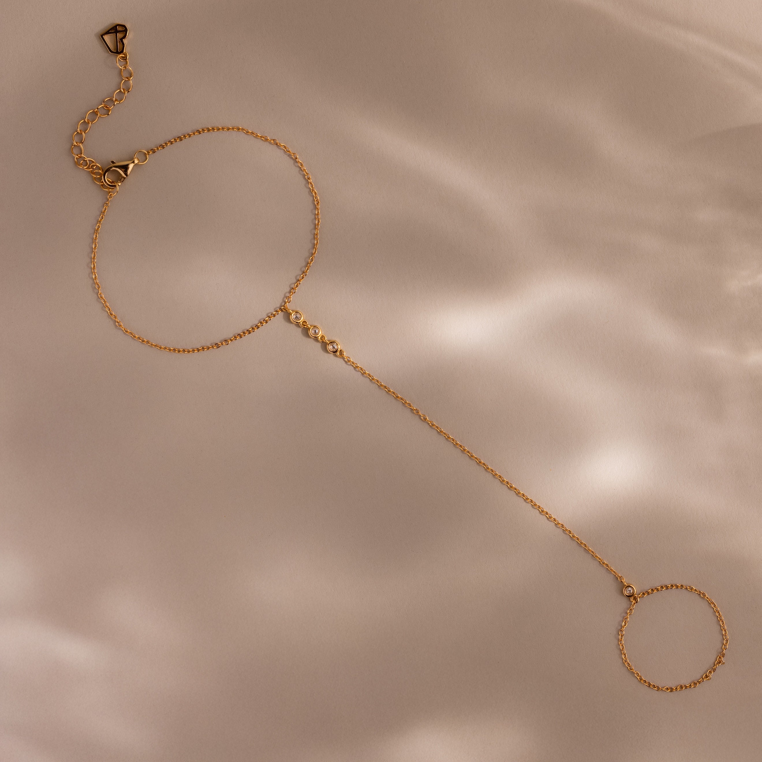 A gold hand chain bracelet with delicate diamonds laid flat on a beige surface with soft shadows.