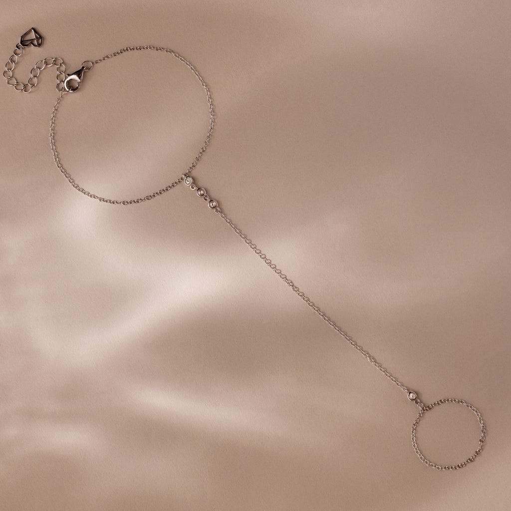 A silver hand chain bracelet with delicate diamonds laid flat on a beige surface with soft shadows.