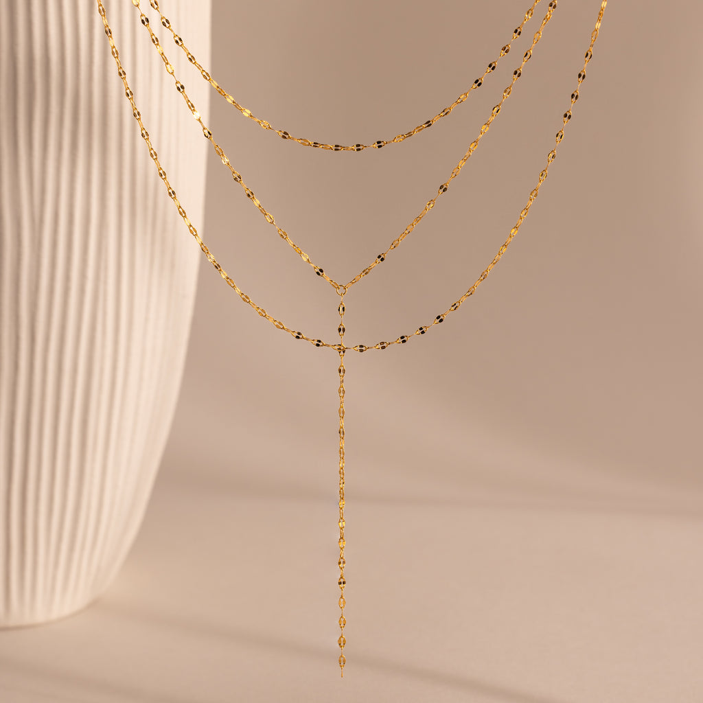 The Sparkling Layered Lariat Necklace in 18K Gold features delicate chains, perfect for effortless layering and summer style, shown against a neutral background with a textured vase.