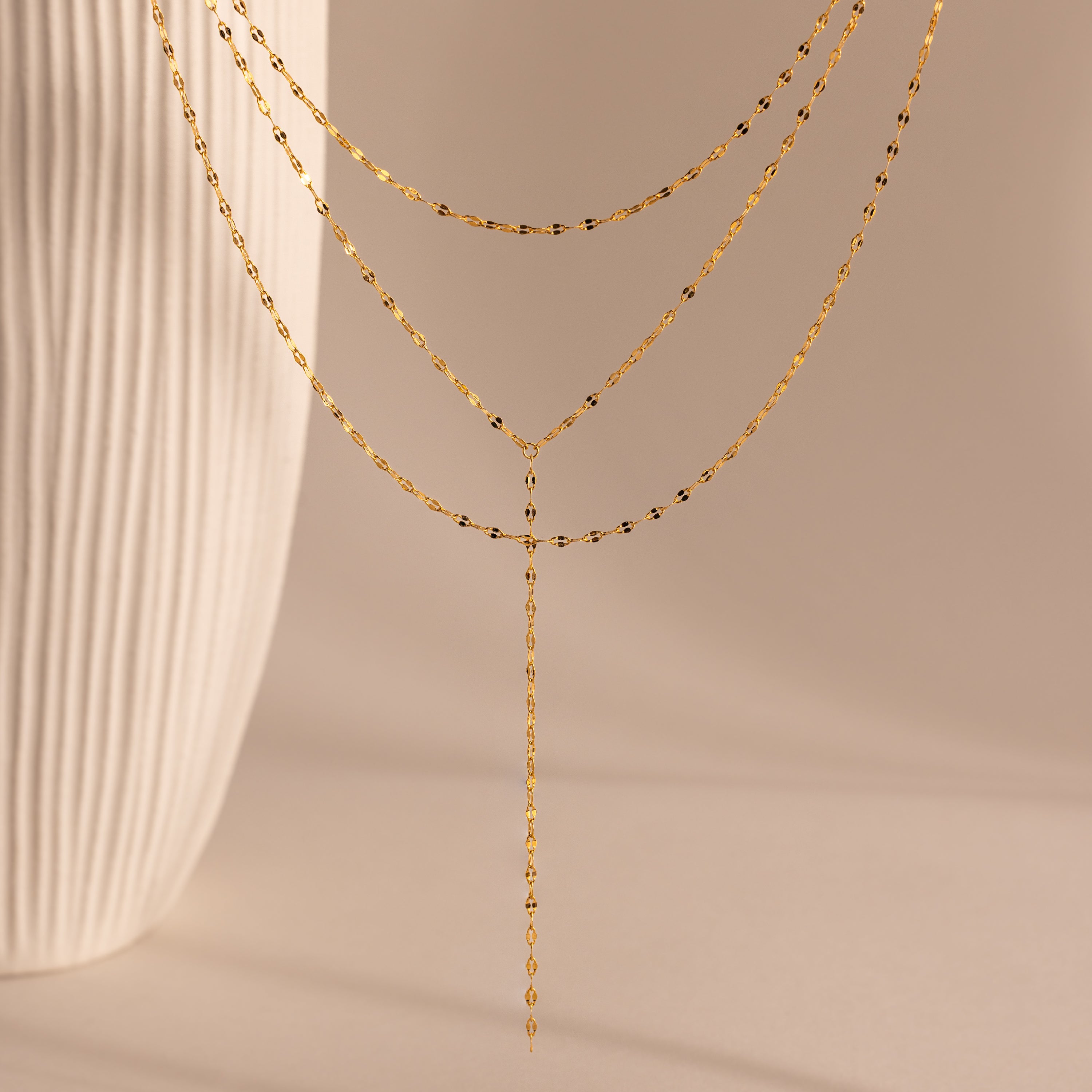 The Sparkling Layered Lariat Necklace in 18K Gold features delicate chains, perfect for effortless layering and summer style, shown against a neutral background with a textured vase.