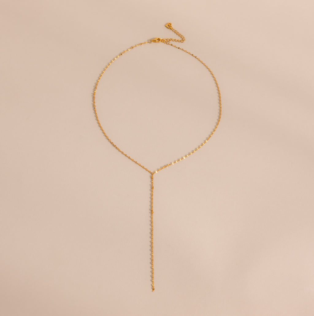 The Mirror Chain Lariat Necklace features a delicate gold chain with a drop pendant and reflective finish, displayed on a light beige background.