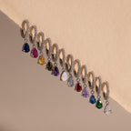 Twelve Pave Birthstone Drop Huggies, featuring silver hoops with pavé diamonds and colorful gemstone drops, are arranged in a row on a beige surface.