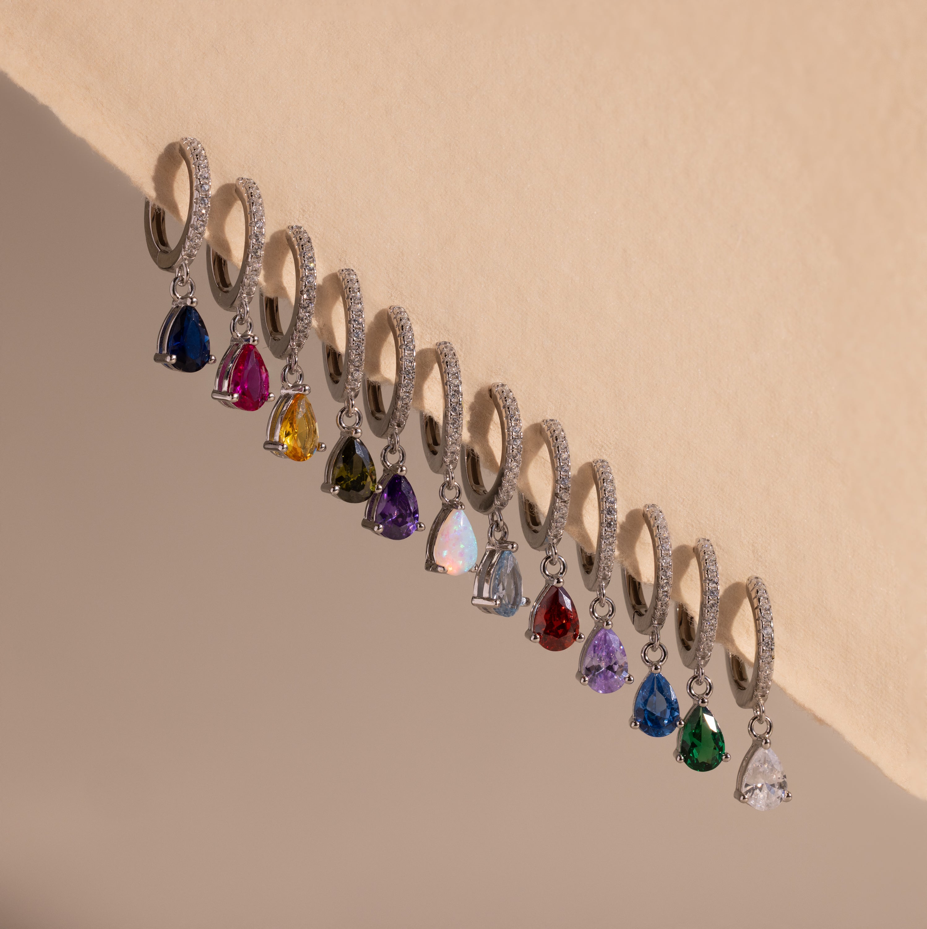 Twelve Pave Birthstone Drop Huggies, featuring silver hoops with pavé diamonds and colorful gemstone drops, are arranged in a row on a beige surface.