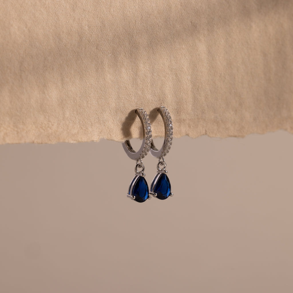 Pave Birthstone Drop Huggies featuring silver hoops and blue teardrop gemstones hang from a textured beige surface, offering an elegant look inspired by classic pavé diamond hoop designs.