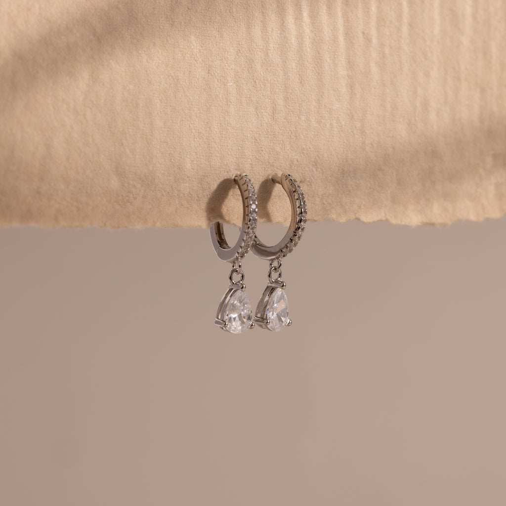 Diamond Drop Huggies in Sterling Silver feature teardrop-shaped crystals hanging from silver hoops, displayed on textured beige paper—an elegant accessory to elevate any semi-formal look against a neutral background.