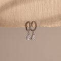 Diamond Drop Huggies in Sterling Silver feature teardrop-shaped crystals hanging from silver hoops, displayed on textured beige paper—an elegant accessory to elevate any semi-formal look against a neutral background.
