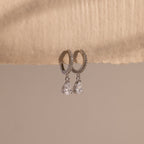 Diamond Drop Huggies in Sterling Silver feature teardrop-shaped crystals hanging from silver hoops, displayed on textured beige paper—an elegant accessory to elevate any semi-formal look against a neutral background.