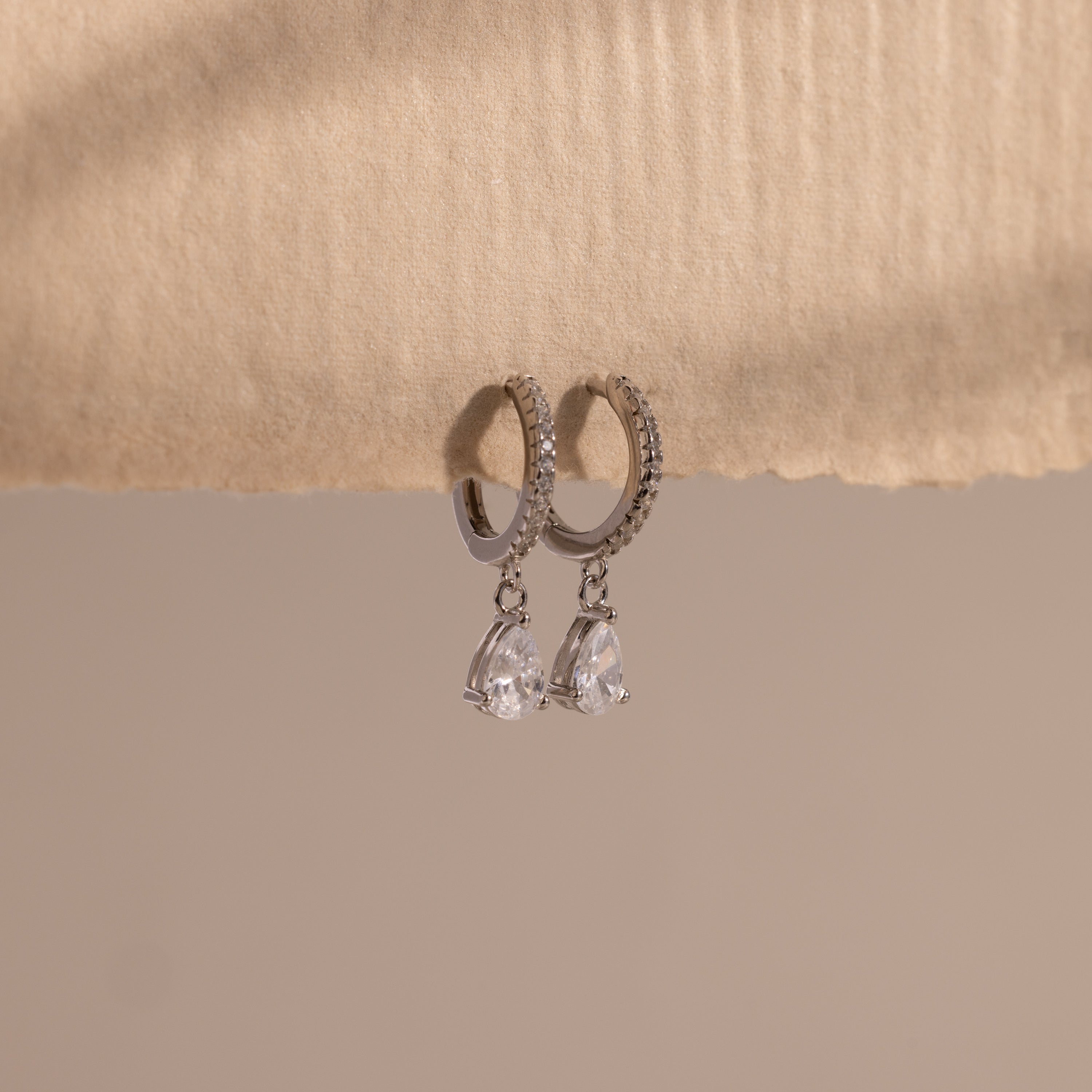 Diamond Drop Huggies in Sterling Silver feature teardrop-shaped crystals hanging from silver hoops, displayed on textured beige paper—an elegant accessory to elevate any semi-formal look against a neutral background.