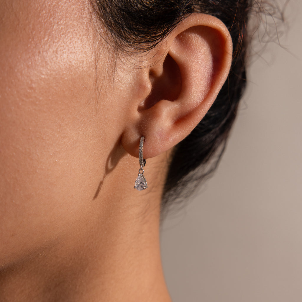 Close-up of a person’s ear wearing Diamond Drop Huggies in Sterling Silver—a small, diamond-encrusted hoop with a dangling teardrop gem—ideal for adding elegance to semi-formal looks.